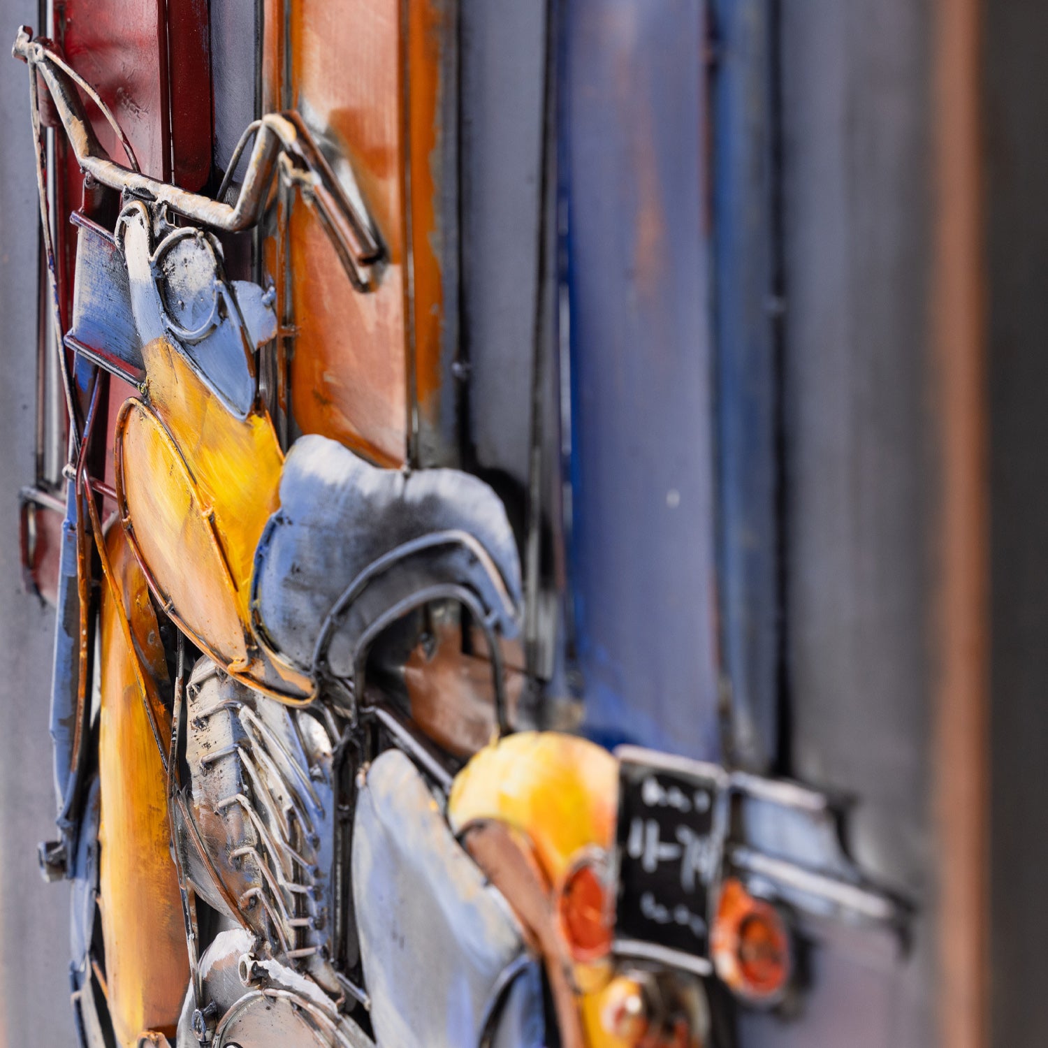 Close-up of a colorful metal wall art sculpture depicting stylized vintage motorbikes and mechanical parts in blue, yellow, orange, and silver tones.