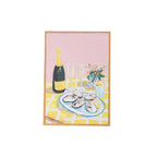 A framed still life painting featuring a bottle of champagne, two glasses, a plate of oysters, and a small flower arrangement on a yellow-and-white checkered table against a pink background, shown on a white backdrop.