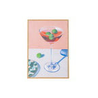 A framed still life painting featuring a martini glass with green olives in a pink drink, set against a split pink and white background with a blue shadow, enclosed in a light wood frame.