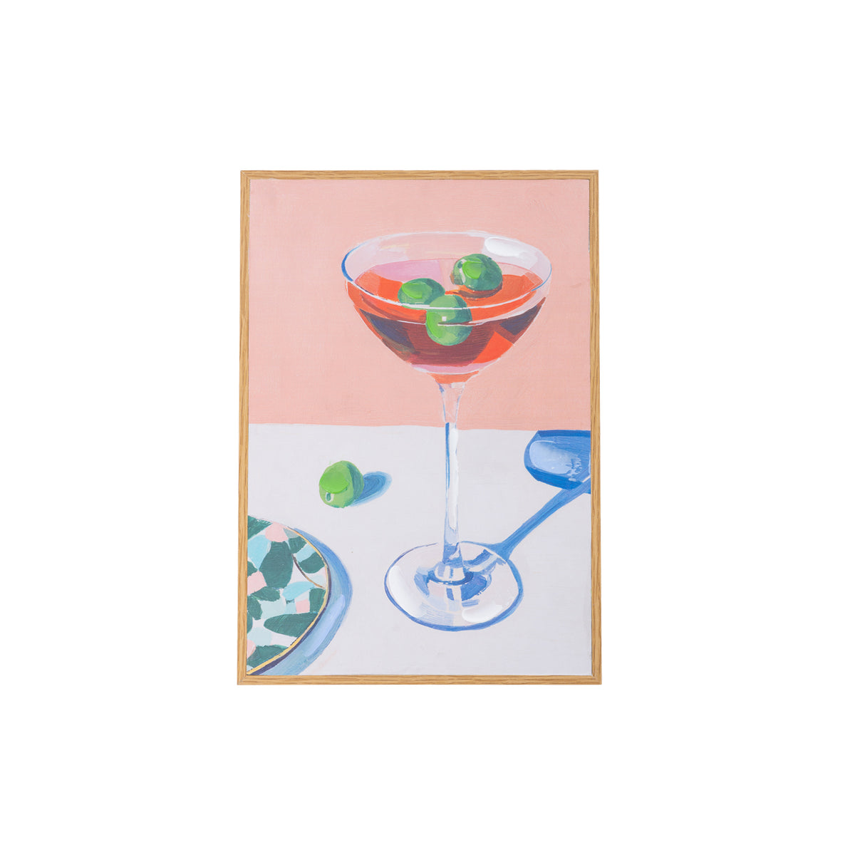 A framed still life painting featuring a martini glass with green olives in a pink drink, set against a split pink and white background with a blue shadow, enclosed in a light wood frame.