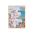 A framed art print depicting a colorful Mediterranean coastal village scene with white buildings, turquoise water, and lush tropical foliage, shown against a plain white background.