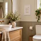 A styled bathroom interior with a white vessel sink, marble countertop, wooden vanity, framed wall art reading 'Rain down the global vibes,' wainscoting, a toilet, and decorative flowers and greenery.