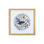 Square wall art with a natural wood-look frame, featuring a blue and brown Piwakawaka with its distinctive fanned tail, perched amongst delicate Kowhai flowers and botanical illustrations in a circular design on a white background.
