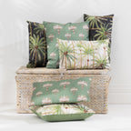 A set of tropical palm tree patterned outdoor cushions in sage green, cream, and black, styled on and around a woven wicker storage trunk against a white wall.