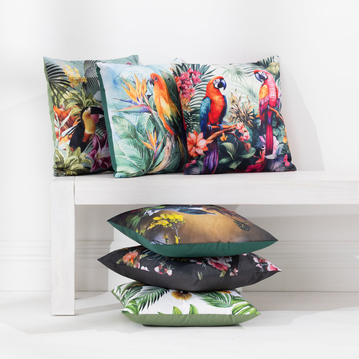 A collection of outdoor cushions with various colorful tropical and nature prints, arranged on and beside a white wooden bench against a white wall.