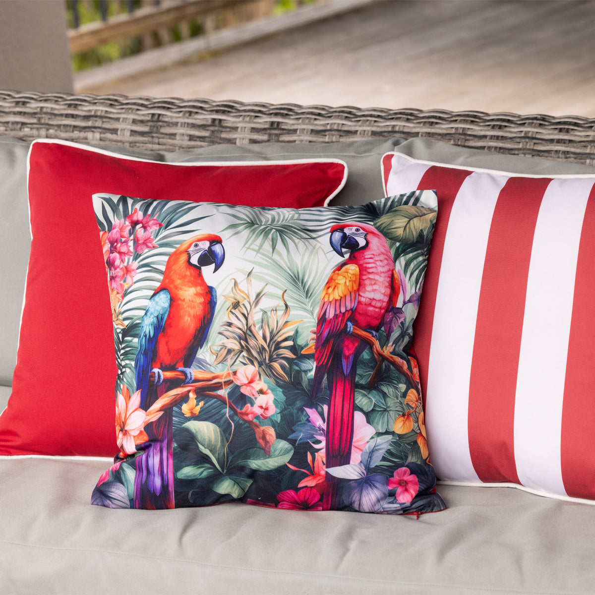 An outdoor cushion featuring a colorful print of two macaws in a tropical jungle setting, styled on a gray outdoor sofa alongside red and red-and-white striped cushions.