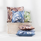 A collection of outdoor cushions with tropical leaf patterns in blue, green, and brown tones, arranged around a woven storage basket against a clean white wall.