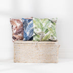 A collection of outdoor cushions with tropical leaf patterns in blue, green, and brown tones, arranged around a woven storage basket against a clean white wall.