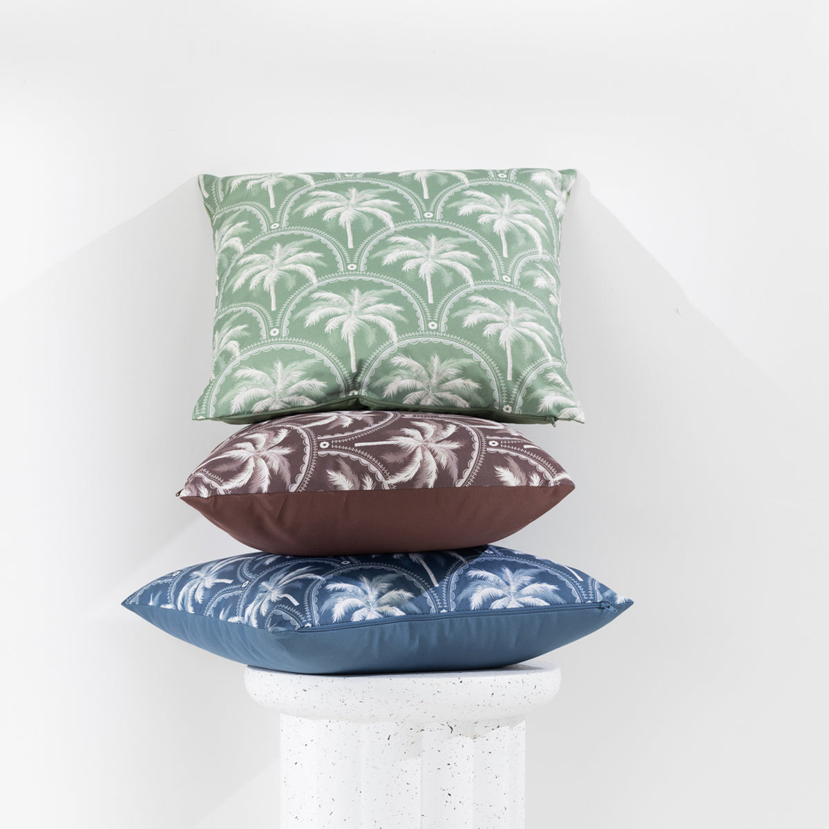 A collection of outdoor cushions with tropical palm tree design in blue, green, and brown tones, arranged around a side table against a clean white wall.