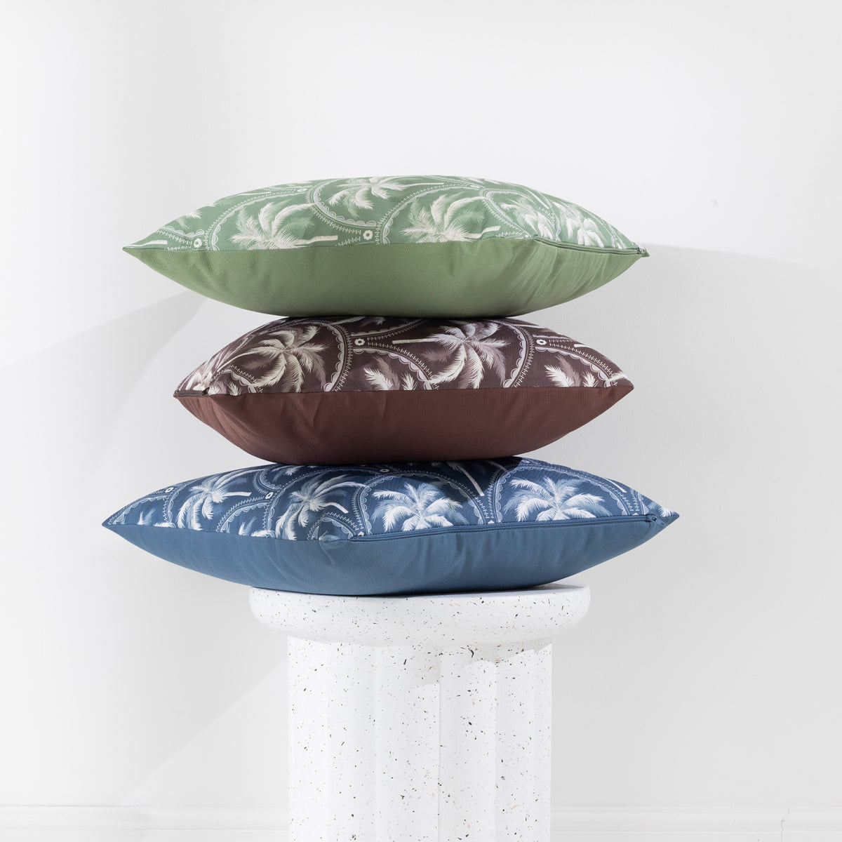 A collection of outdoor cushions with tropical palm tree design in blue, green, and brown tones, arranged around a side table against a clean white wall.
