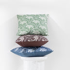 A collection of outdoor cushions with tropical palm tree design in blue, green, and brown tones, arranged around a side table against a clean white wall.