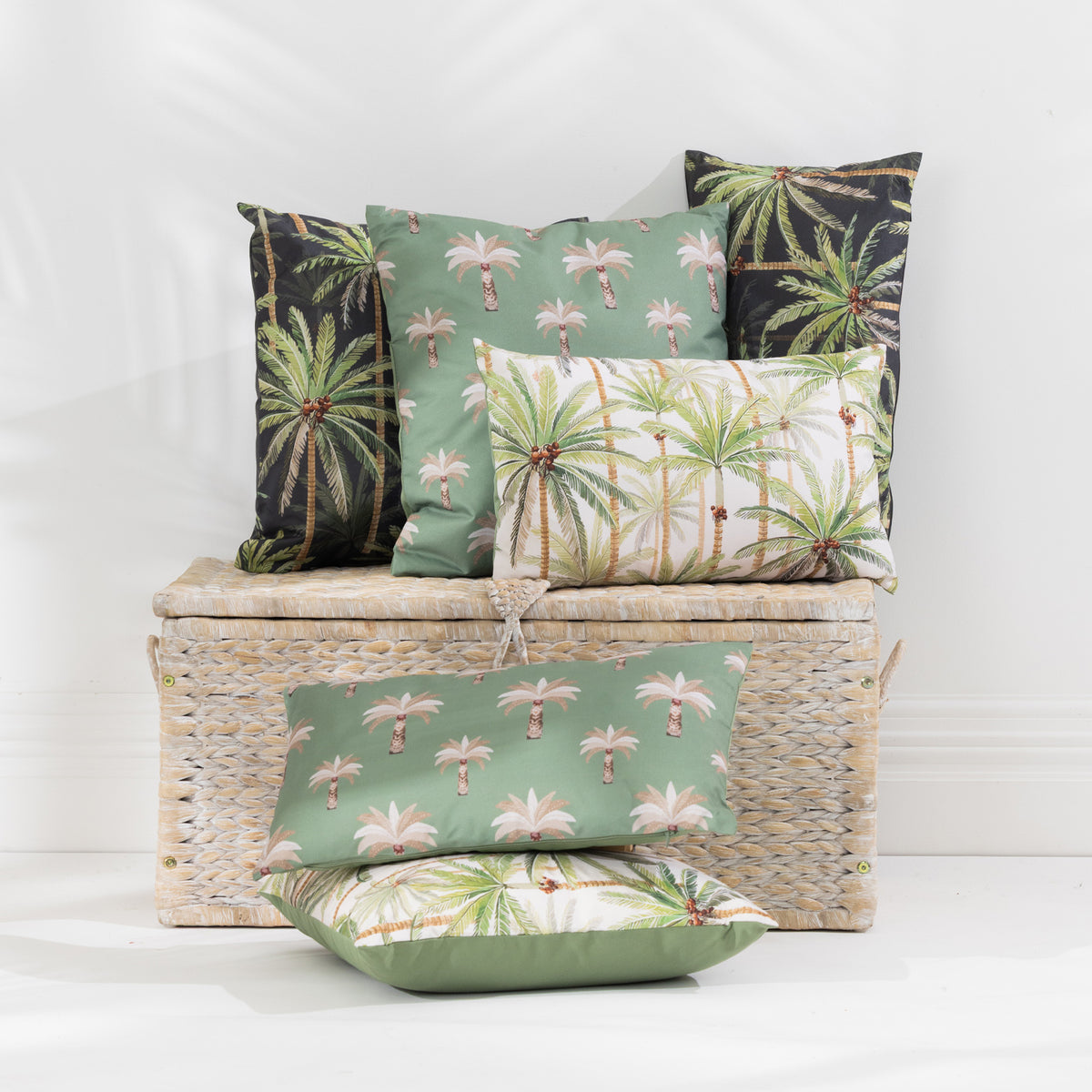 A set of tropical palm tree patterned outdoor cushions in sage green, cream, and black, styled on and around a woven wicker storage trunk against a white wall.