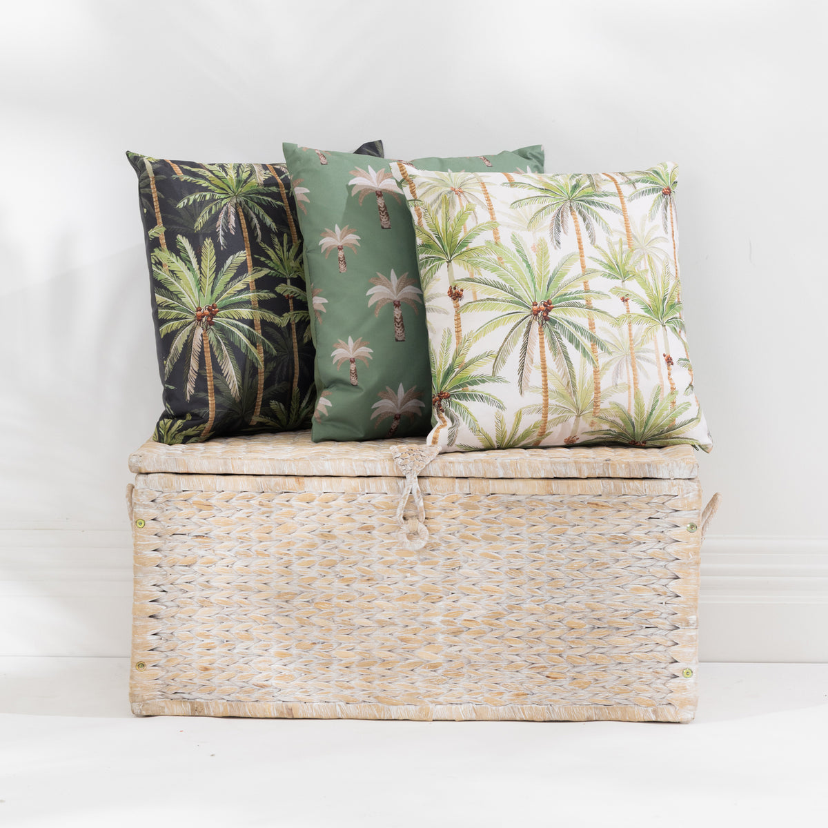 A set of tropical palm tree patterned outdoor cushions in sage green, cream, and black, styled on and around a woven wicker storage trunk against a white wall.