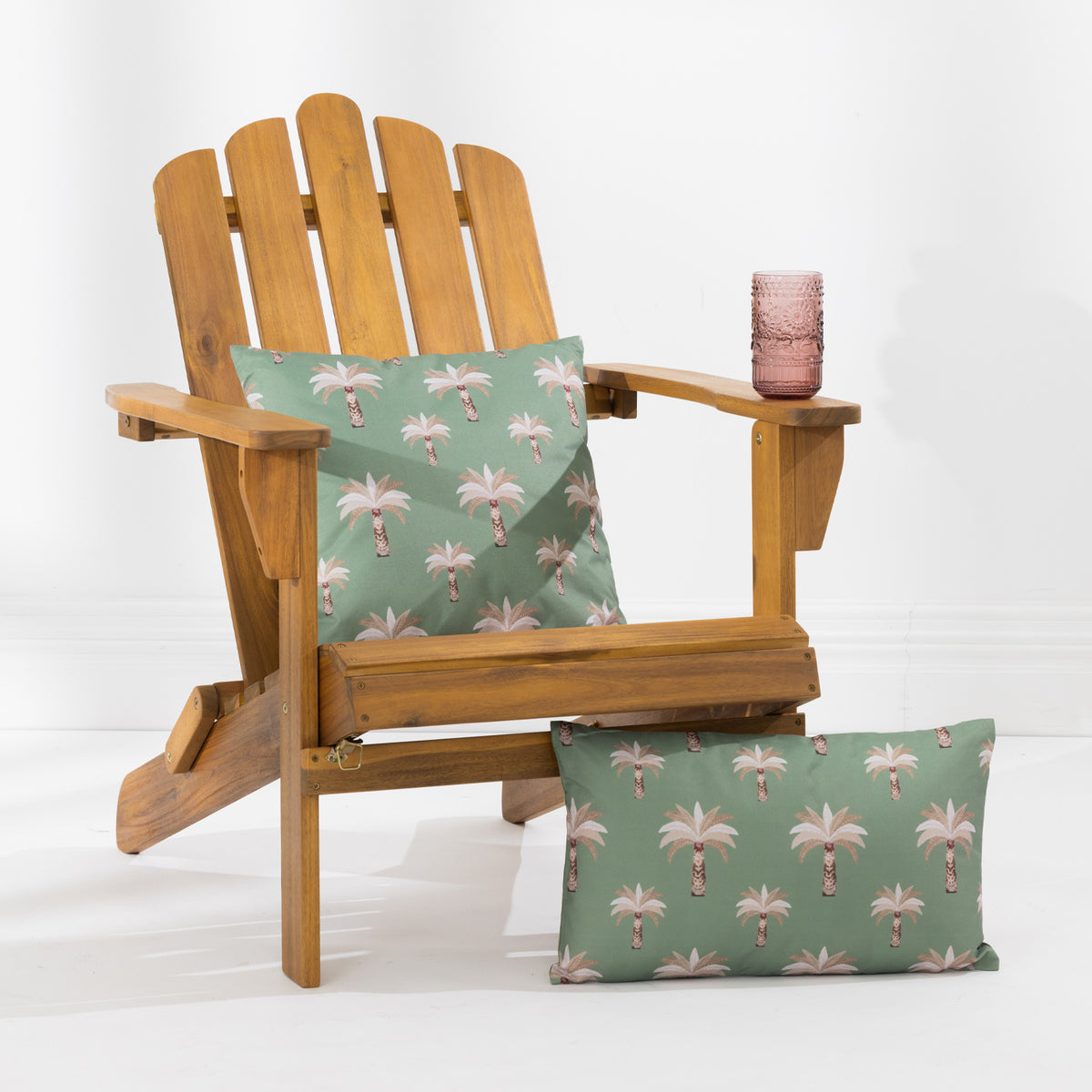 A wooden Adirondack-style chair with two decorative pillows featuring a palm tree pattern on a green background, set against a clean white studio backdrop.