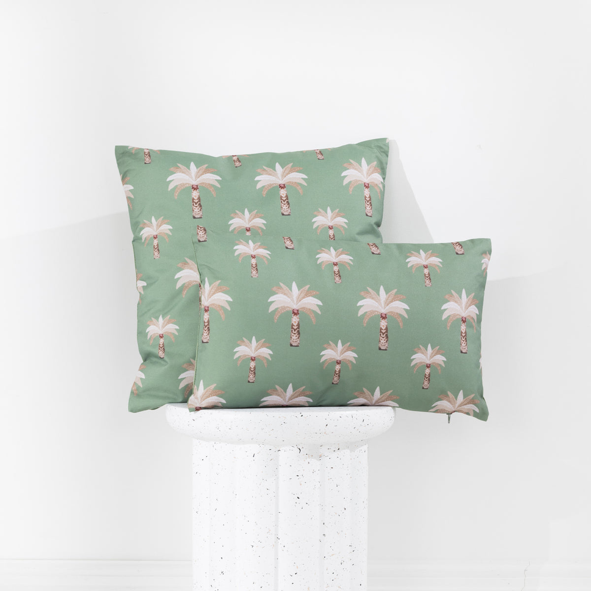 Two outdoor cushions with a green background and a repeating palm tree pattern in white, displayed on a white speckled pedestal against a white wall.