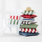 A stack of colorful striped and solid outdoor cushions sits beside a white Adirondack chair in a bright studio setting. A straw hat rests on top of the cushions.