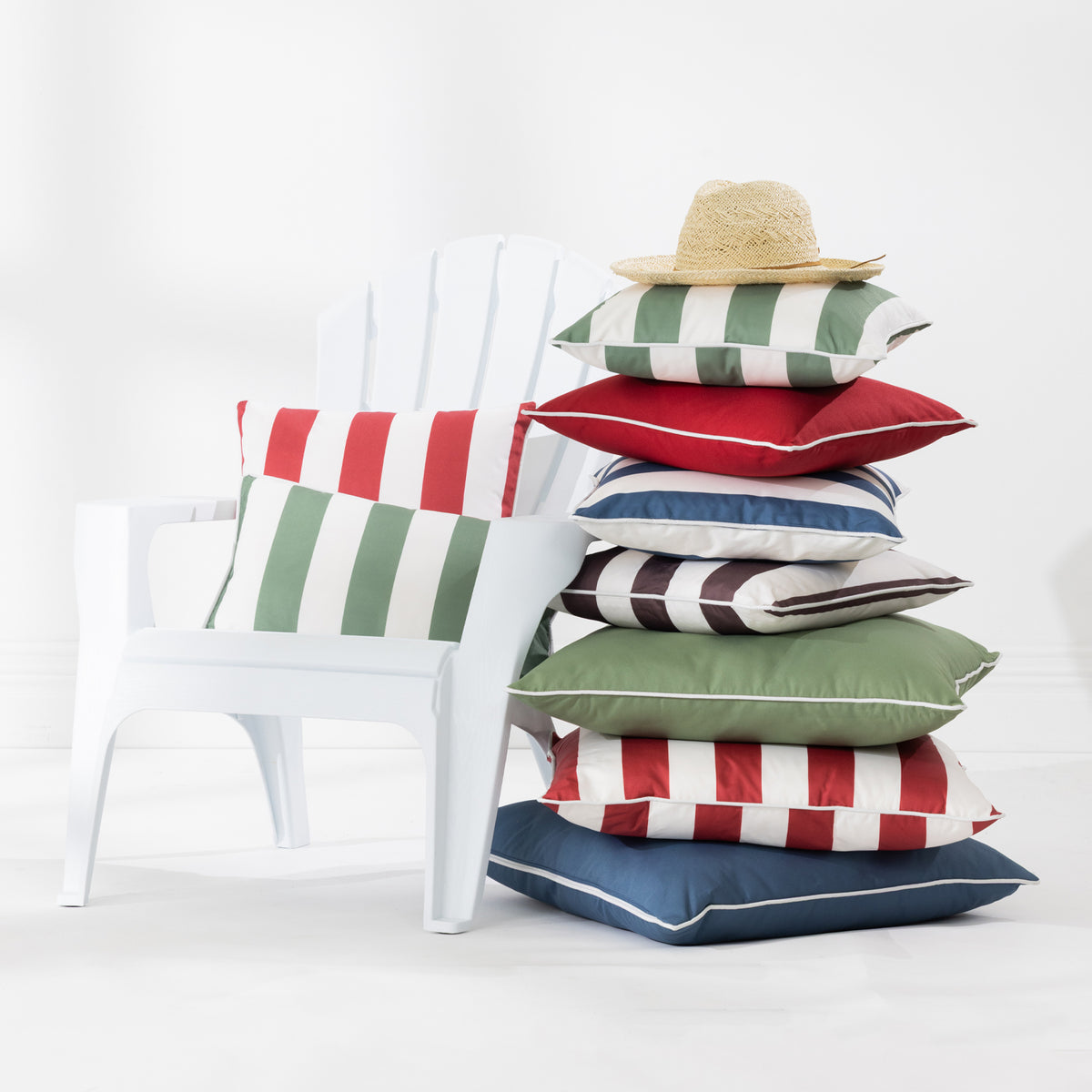 A stack of colorful striped and solid outdoor cushions sits beside a white Adirondack chair in a bright studio setting. A straw hat rests on top of the cushions.