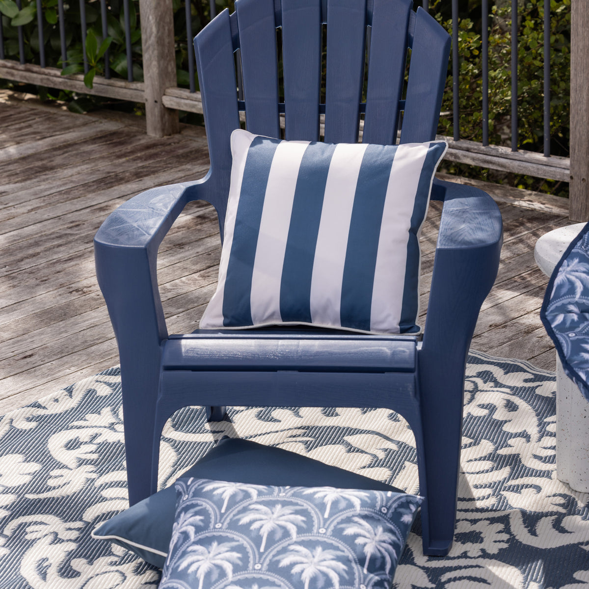 A blue outdoor Adirondack chair on a patterned rug with matching blue and white cushions, set on a wooden deck with a railing and greenery in the background.
