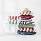 A stack of colorful striped outdoor cushions sits beside a white Adirondack chair in a bright studio setting. A straw hat rests on top of the cushions.