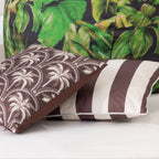 Two outdoor cushions arranged on a white surface: one with a mocha brown and white palm tree pattern, one with mocha brown and white vertical stripes, displayed in front of a tropical green bean bag in the background.