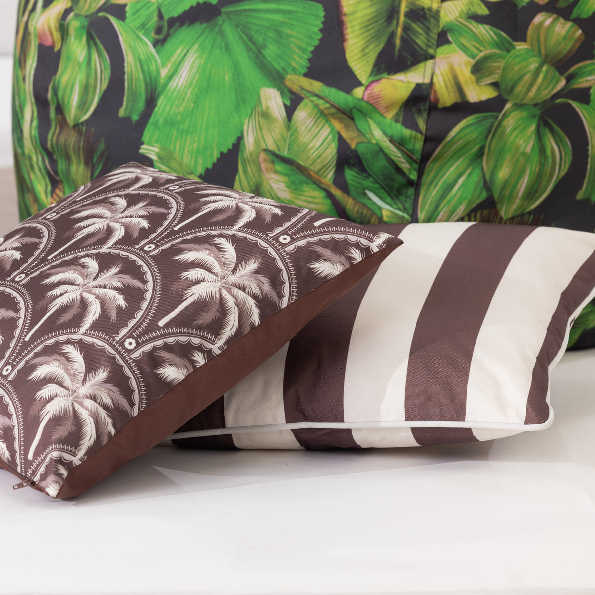 Two outdoor cushions arranged on a white surface: one with a mocha brown and white palm tree pattern, one with mocha brown and white vertical stripes, displayed in front of a tropical green bean bag in the background.