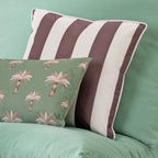 A decorative outdoor cushion with vertical mocha brown and off-white stripes sits on a green sofa, styled with a smaller green pillow featuring a palm tree pattern.