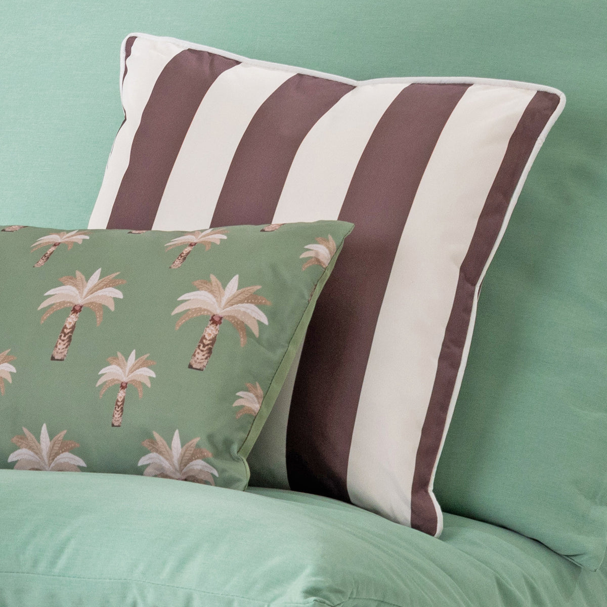 A decorative outdoor cushion with vertical mocha brown and off-white stripes sits on a green sofa, styled with a smaller green pillow featuring a palm tree pattern.