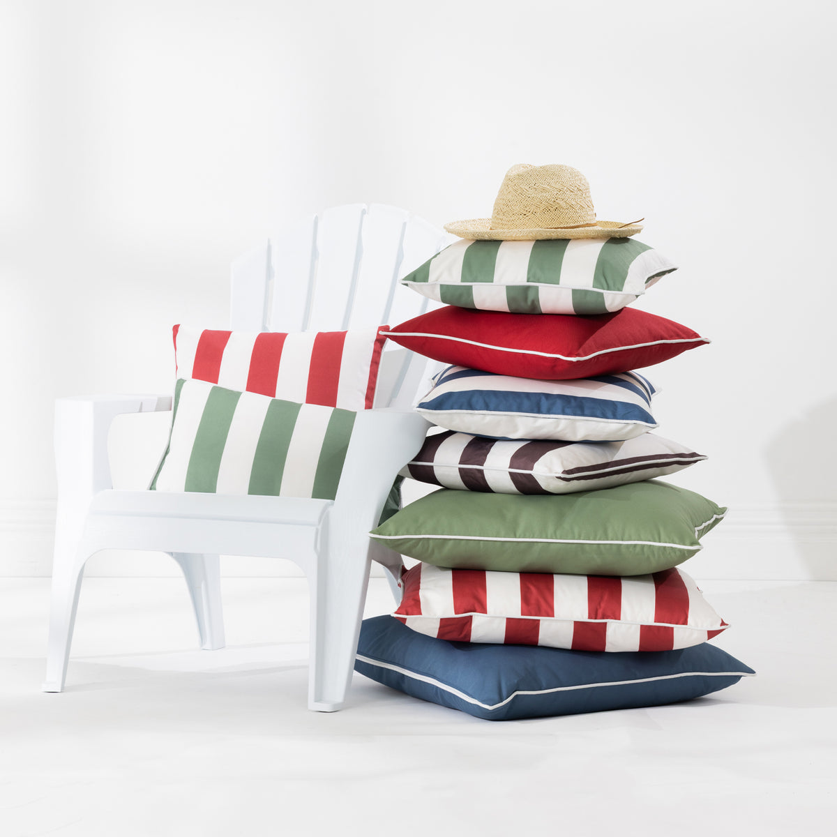 A stack of colorful striped outdoor cushions sits beside a white Adirondack chair in a bright studio setting. A straw hat rests on top of the cushions.