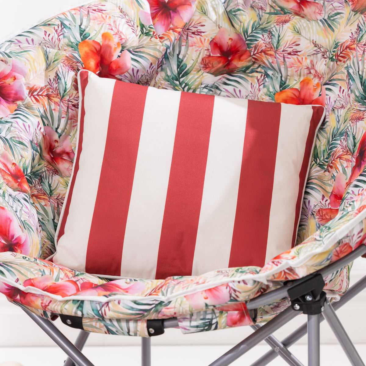 An outdoor cushion with vertical red and white stripes sits on a tropical floral moon chair with gray metal legs.