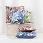 Four outdoor cushions with tropical leaf patterns in various colors, styled on and around a woven wicker storage basket against a white wall.