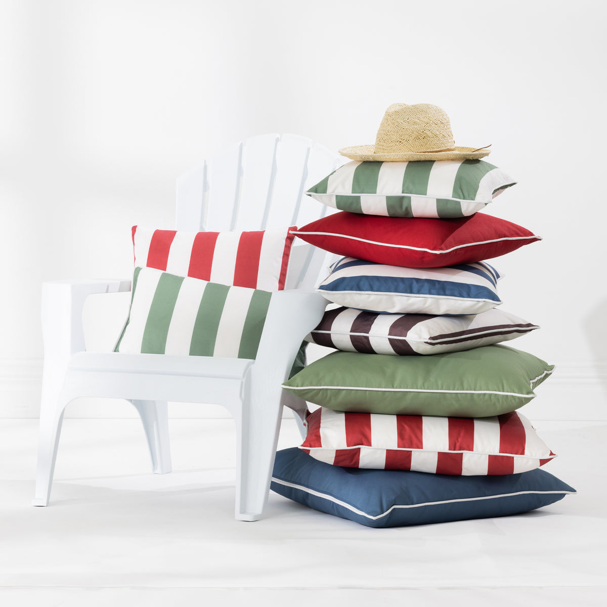A stack of colorful striped and solid outdoor cushions sits beside a white Adirondack chair in a bright studio setting. A straw hat rests on top of the cushions.
