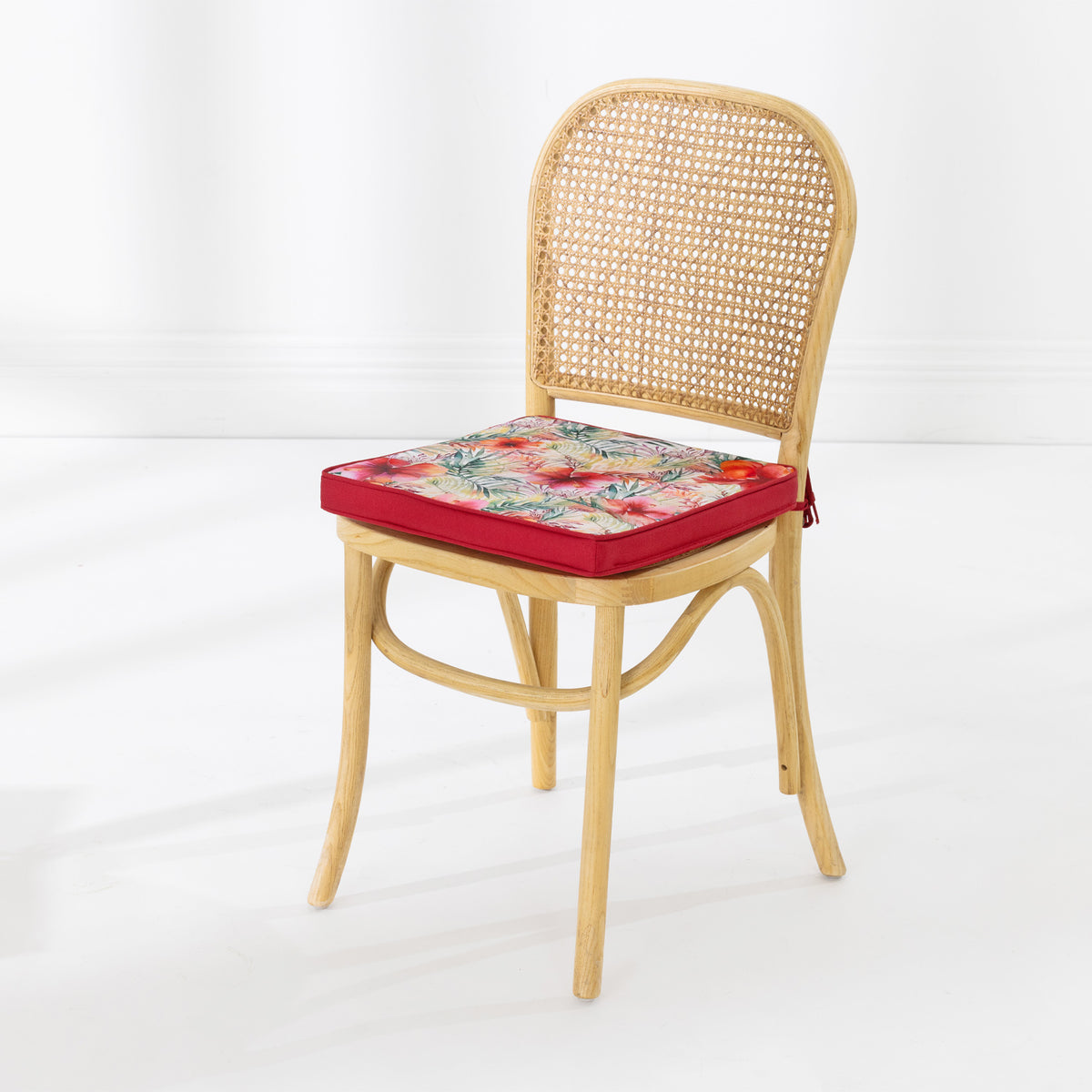 A light natural wood cane dining chair with a red upholstered outdoor seat cushion featuring a colorful floral pattern, set against a clean white studio background.
