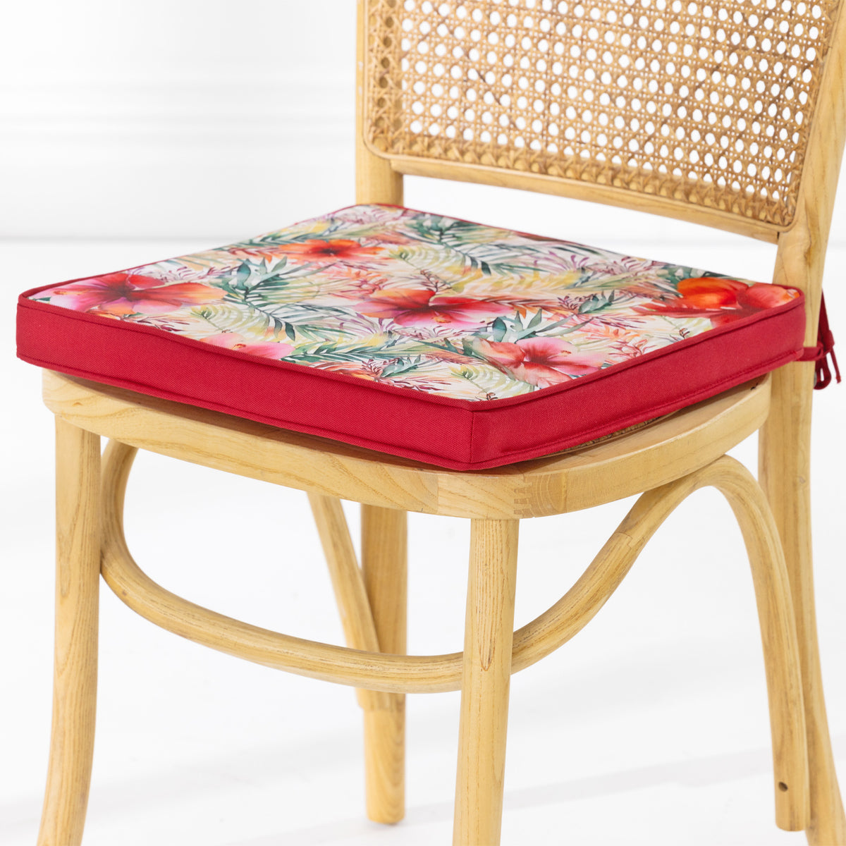 A light natural wood cane dining chair with a red upholstered outdoor seat cushion featuring a colorful floral pattern, set against a clean white studio background.