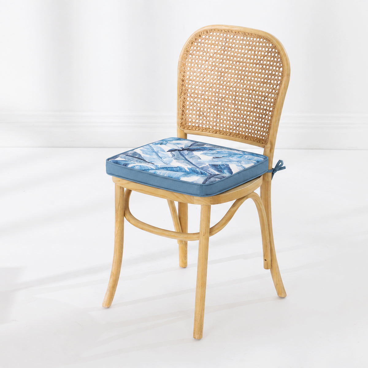A light natural wood dining chair with a woven cane backrest, featuring a blue leaf patterned seat cushion, set against a clean white studio background.