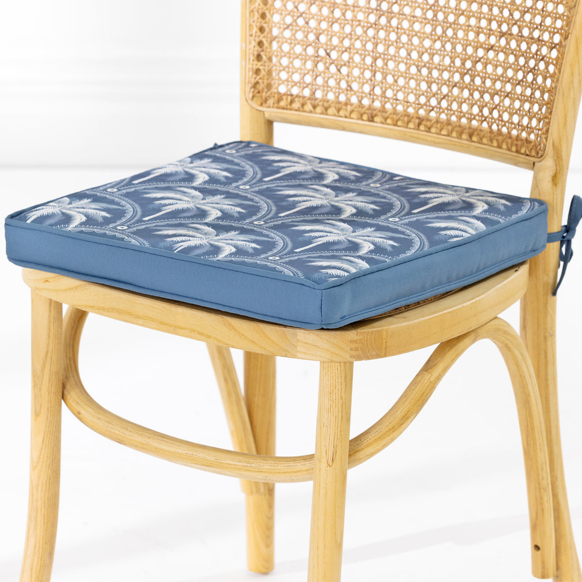 A blue upholstered chair cushion with a white palm tree pattern, shown fitted on a light wood cane dining chair against a white background.