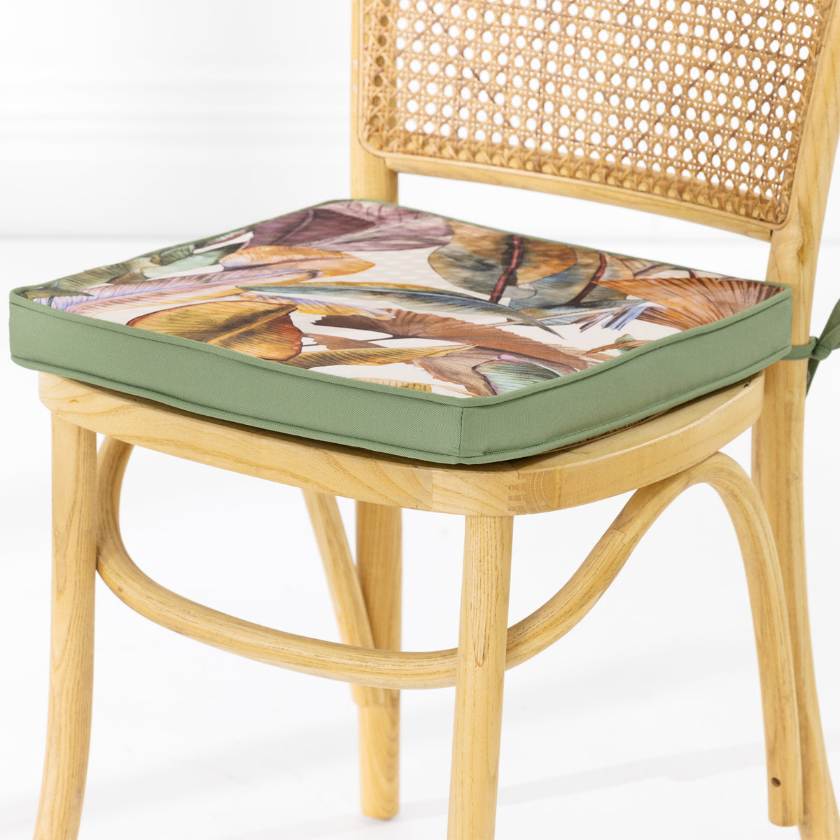 A light natural wood dining chair with a woven cane backrest, featuring a thick seat cushion with a multicolor leaf pattern and an green edge, set against a clean white background.