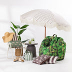 A styled beach and lounge setup on a white studio floor featuring a large white beach umbrella with fringe, a green tropical leaf print bean bag chair, a striped folding chair with a green palm-patterned cushion, a straw hat, a small side table, and matching decorative pillows.