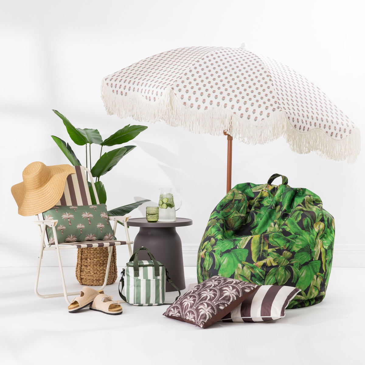 A styled beach and lounge setup on a white studio floor featuring a large white beach umbrella with fringe, a green tropical leaf print bean bag chair, a striped folding chair with a green palm-patterned cushion, a straw hat, a small side table, and matching decorative pillows.