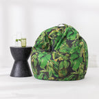 A large bean bag chair with a green tropical leaf print on a black background, styled next to a black speckled side table with a glass mug in a bright, minimalist room.