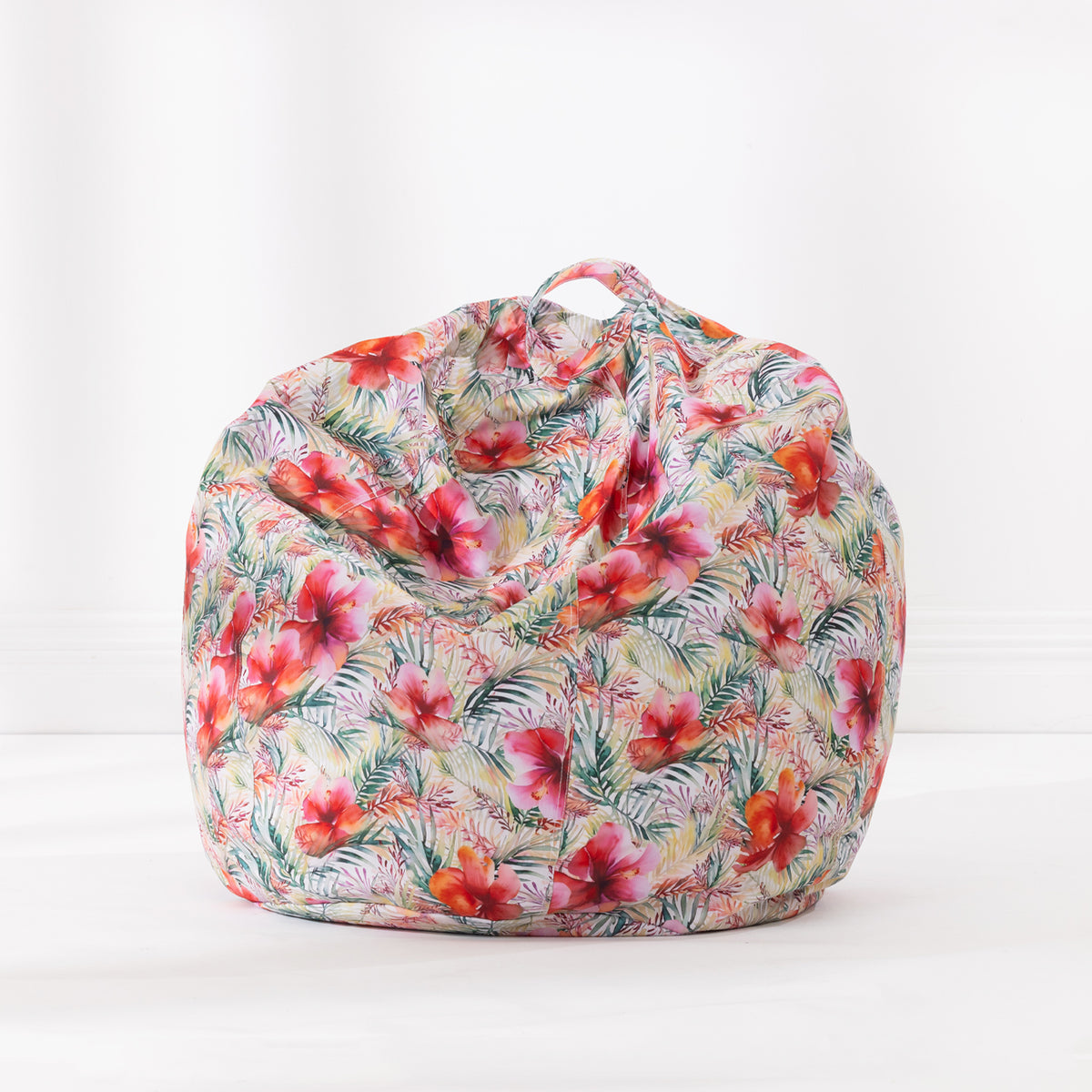 A bean bag chair with a tropical floral print featuring hibiscus flowers and palm leaves in pink, red, green, and orange on a light beige background, set against a plain white studio backdrop.