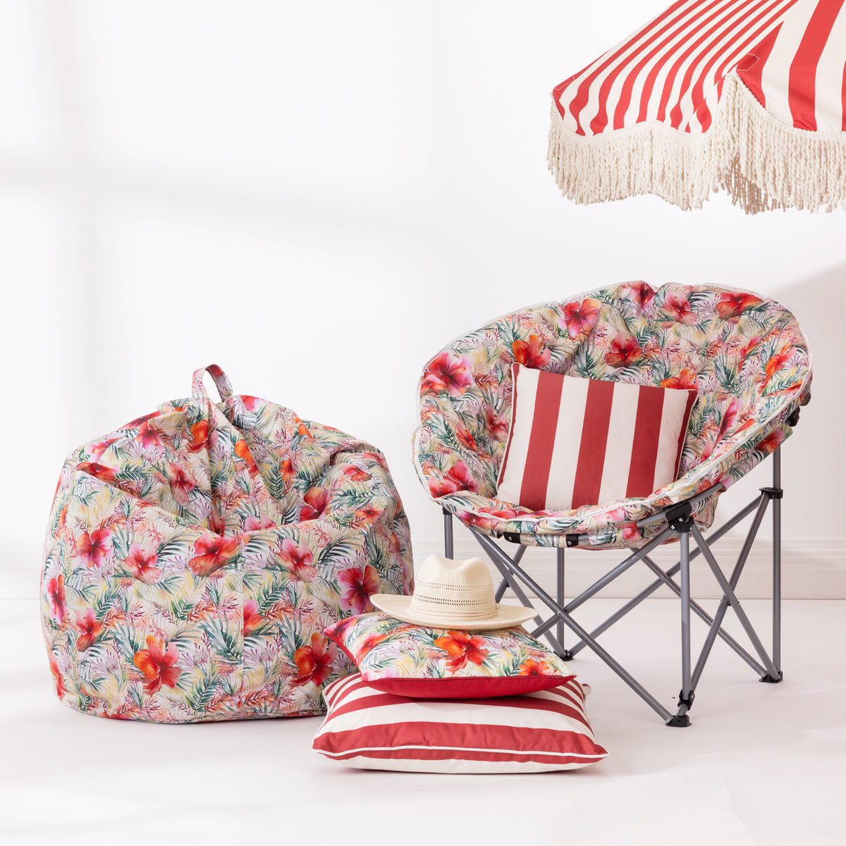 A tropical floral print set displayed on a white studio floor, including a bean bag chair, a folding camp chair with a matching cushion, striped cushions, and a straw hat, with part of a red-and-white striped beach umbrella visible at the top right.