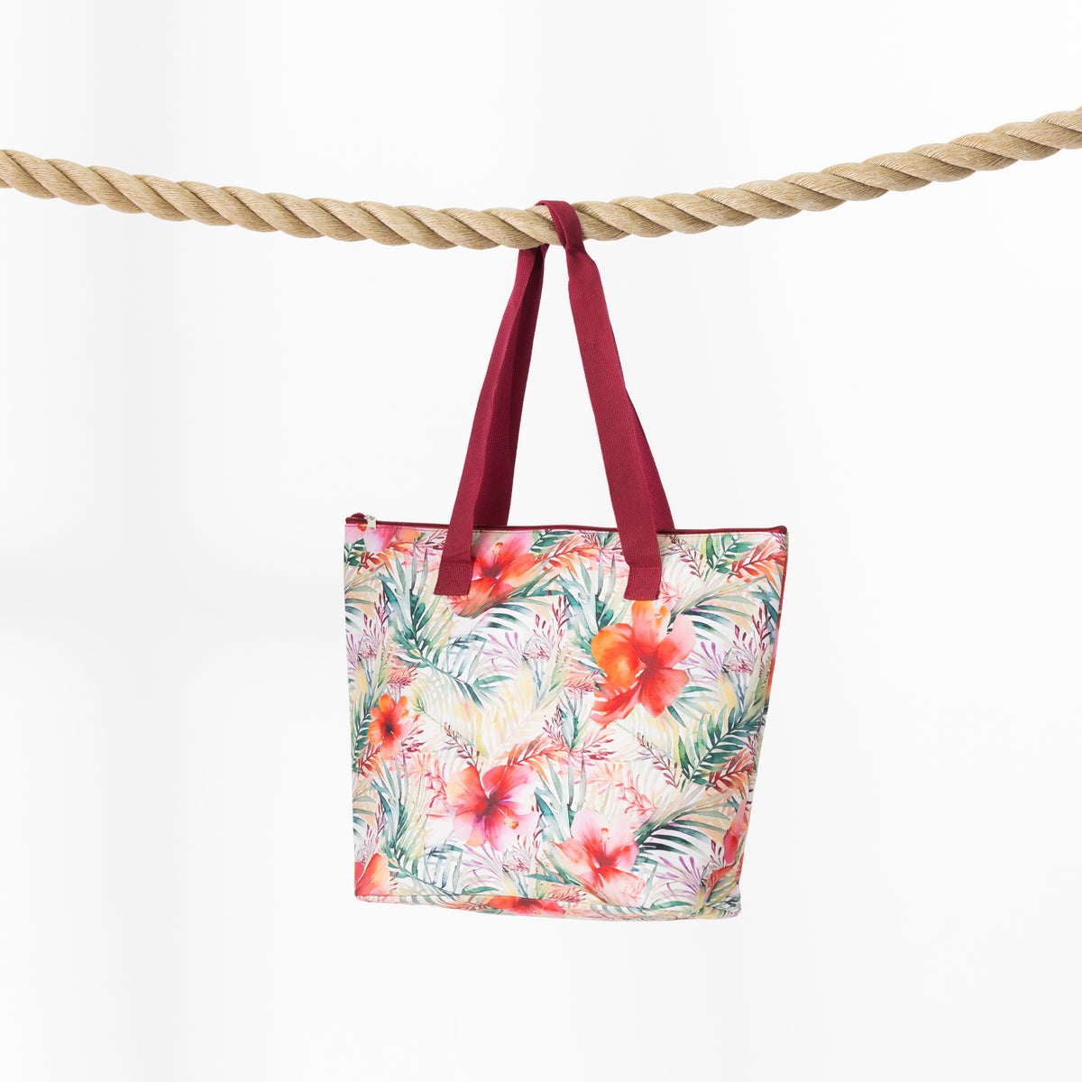 A beach bag with a colorful tropical floral print and dark red handles, hanging from a thick rope against a plain white background.