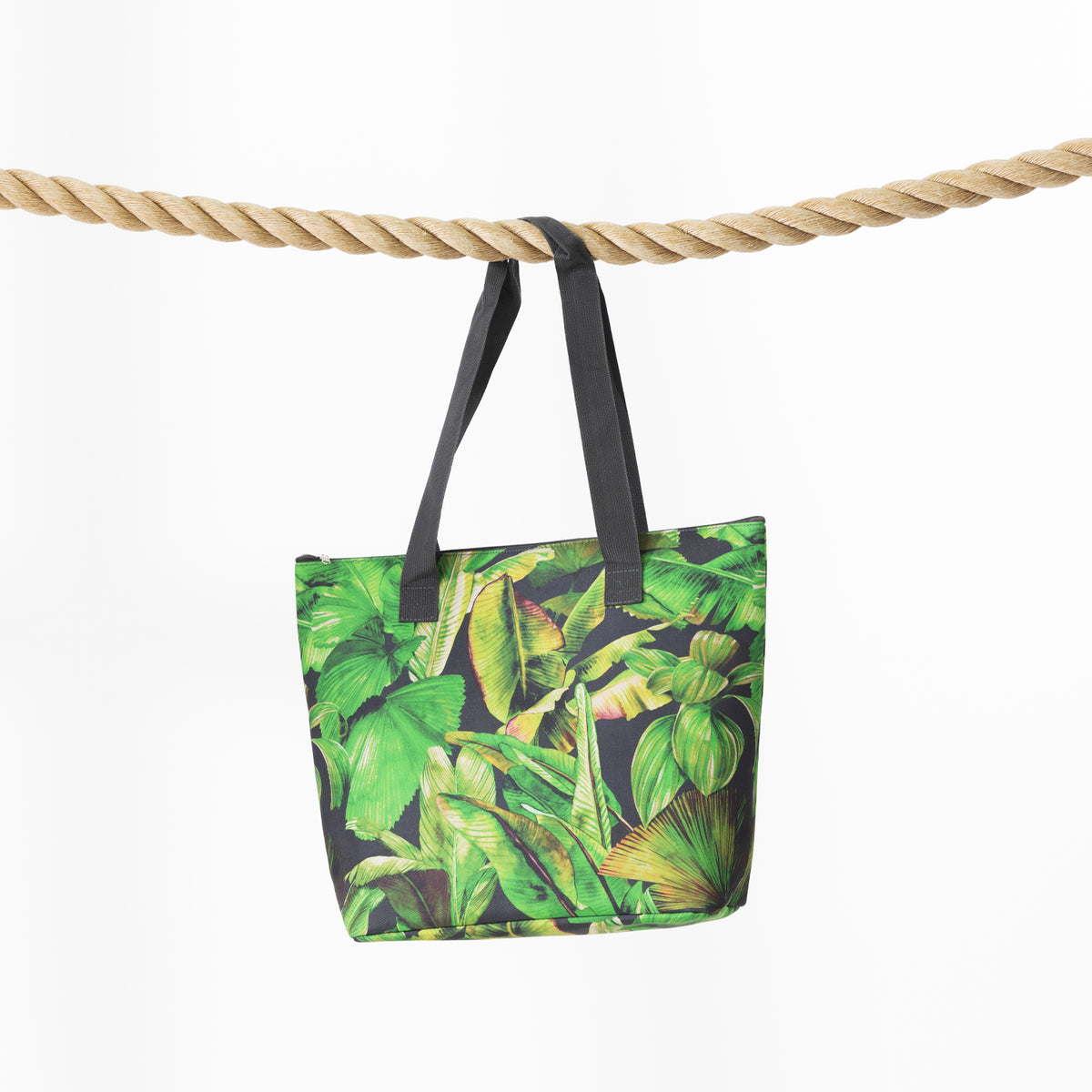A beach bag with a green tropical leaf print and black handles, hanging from a thick natural rope against a plain white background.