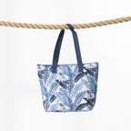 A beach bag with a blue and white tropical leaf print, hanging from a thick natural rope against a plain white background.