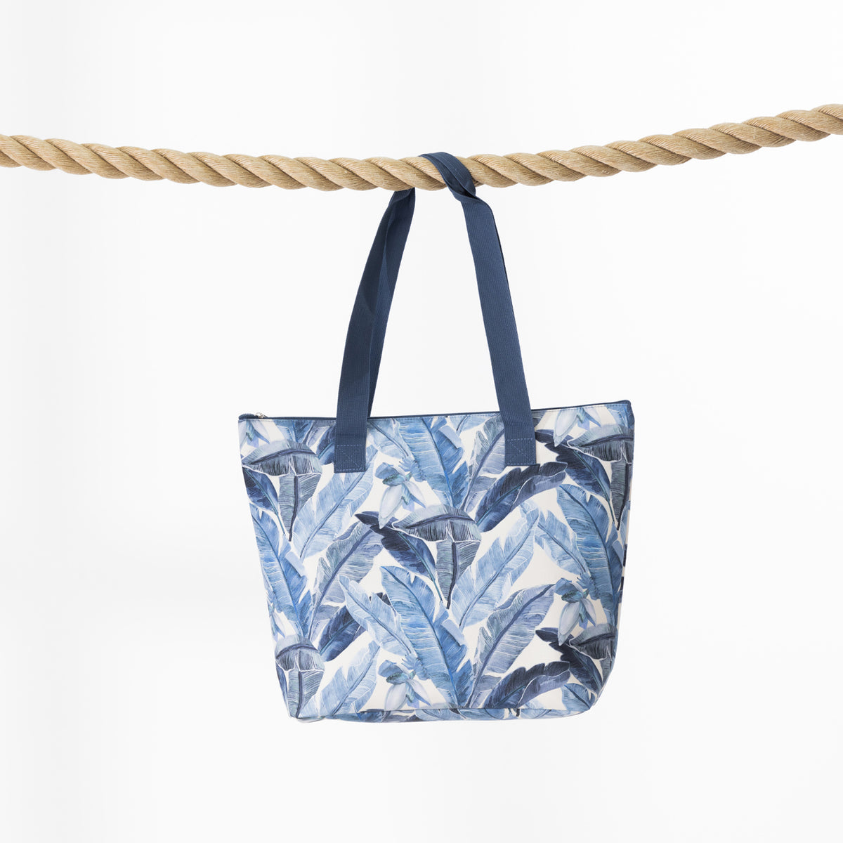 A beach bag with a blue and white tropical leaf print, hanging from a thick natural rope against a plain white background.