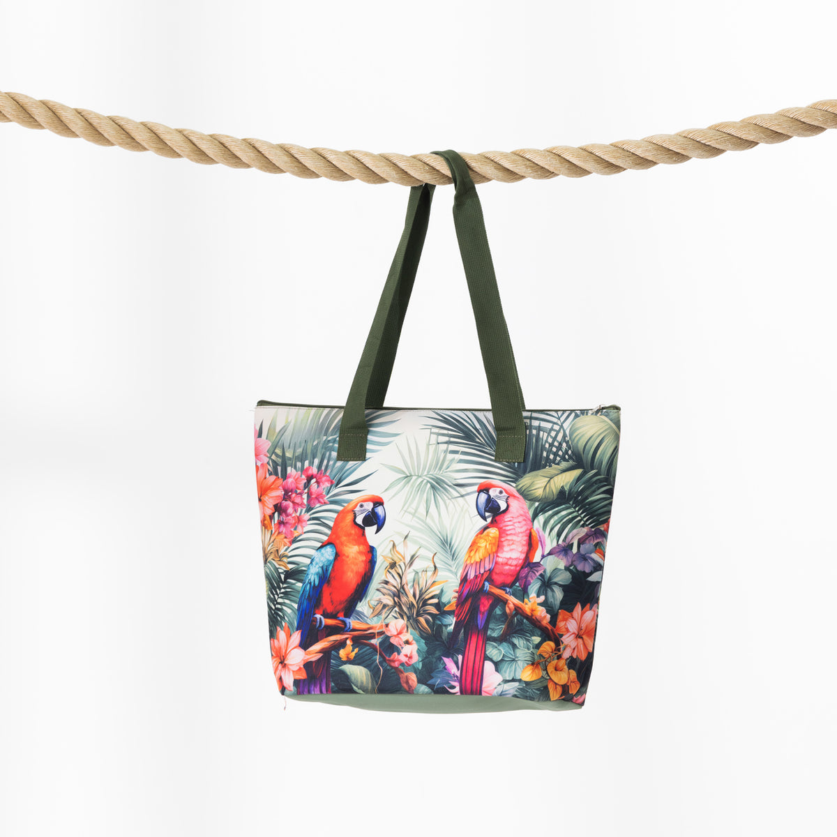 A beach bag with a colorful tropical parrot and palm leaf print, hanging from dark green straps over a thick beige rope against a white background.
