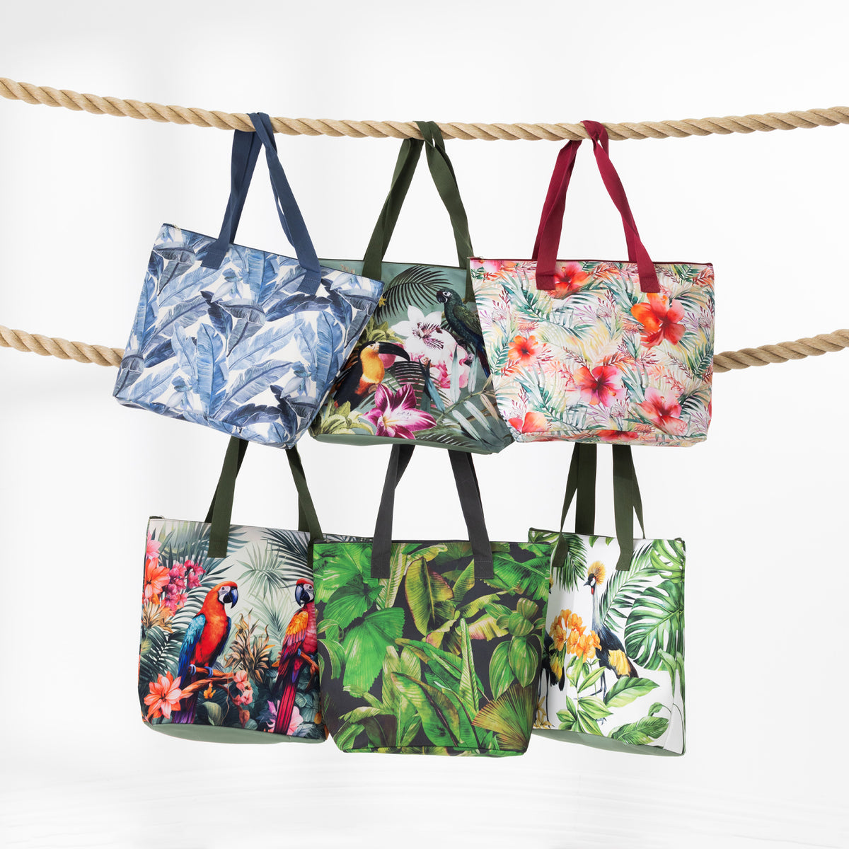 A range of beach bags with tropical and bird prints hanging from a thick rope against a white background.