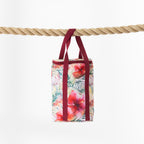 A cooler bag with a tropical floral print hangs from a thick natural rope against a clean white background. The bag features a multicolour pattern of hibiscus flowers and palm leaves in pink, red, green, and yellow, with contrasting dark red handles.