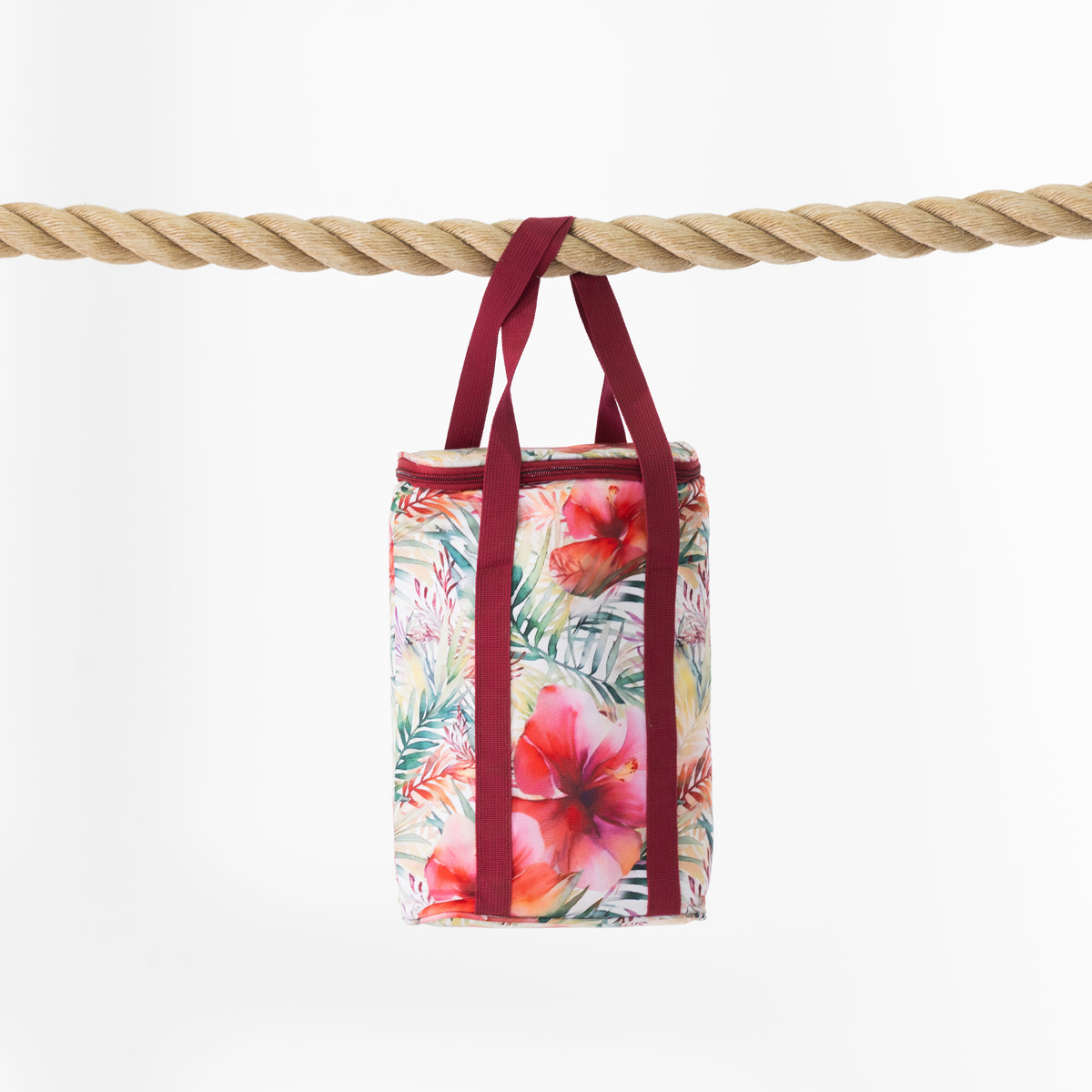 A cooler bag with a tropical floral print hangs from a thick natural rope against a clean white background. The bag features a multicolour pattern of hibiscus flowers and palm leaves in pink, red, green, and yellow, with contrasting dark red handles.