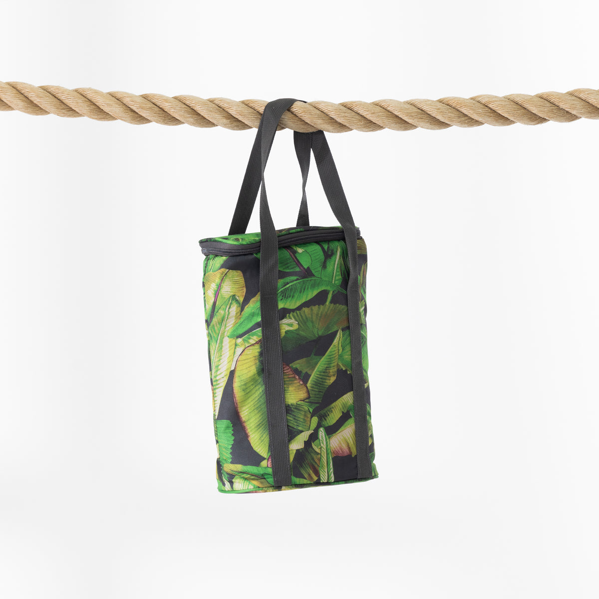 A cooler bag with a green tropical banana leaf pattern and black handles, hanging from a thick natural rope against a clean white background.